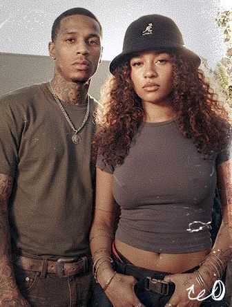 A man and woman stand closely together. The man wears a gray t-shirt and necklace, while the woman wears a hat and cropped top.