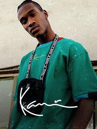 Young man in a green shirt with a black shoulder bag, standing in front of a textured wall with a "Kani" signature overlay.