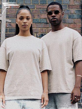 Two people in matching beige outfits stand side by side against a brick wall. One wears sunglasses, and both have a serious expression.