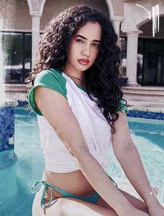 Woman with curly hair in a white and green top sits by a poolside, with a mansion in the background.