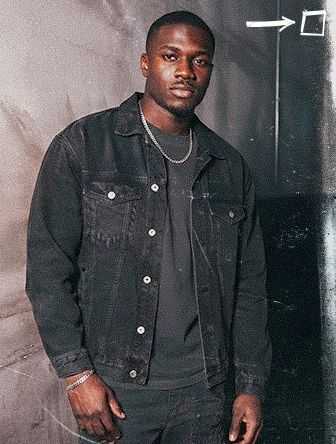 Man in a black denim jacket and chain necklace stands against a textured backdrop with a white arrow pointing towards the top right corner.