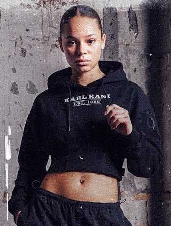 Person wearing a black cropped hoodie and pants stands confidently against a textured wall.