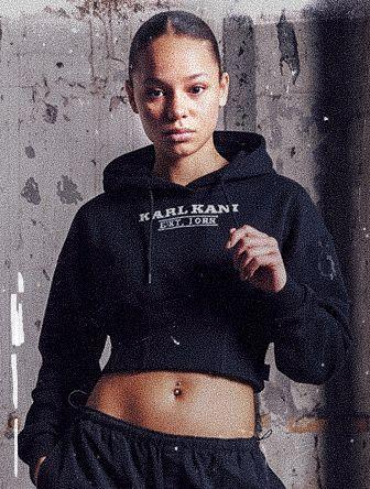 Person wearing a black cropped hoodie and pants stands confidently against a textured wall.