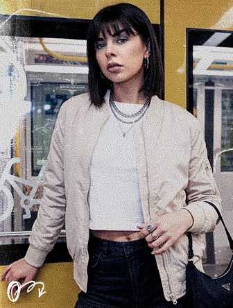 Woman with bangs in a beige jacket and white top stands in a subway, wearing layered necklaces and hoop earrings.