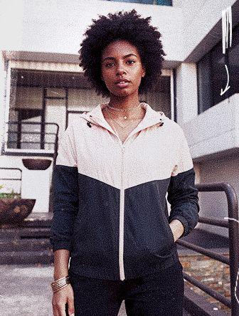 A person with curly hair wearing a two-toned hoodie stands confidently outdoors in front of a modern building.