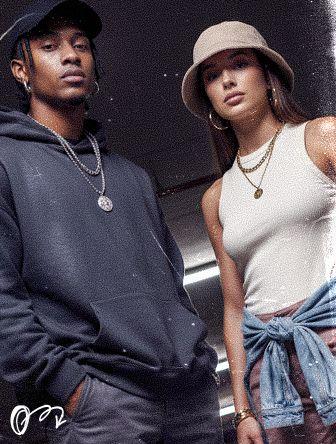 Two stylish individuals, one in a hoodie and cap, the other in a hat and tank top, pose confidently in casual urban attire.