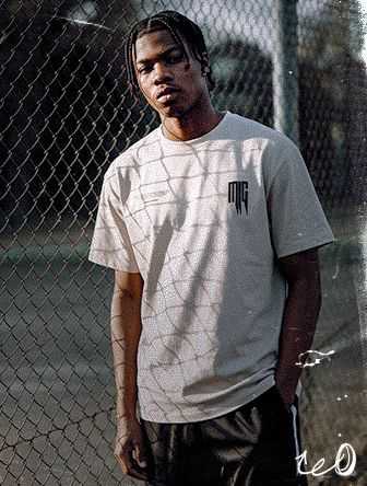 Man standing by a chain-link fence, wearing a light-colored T-shirt and dark pants, with sunlight casting shadows.