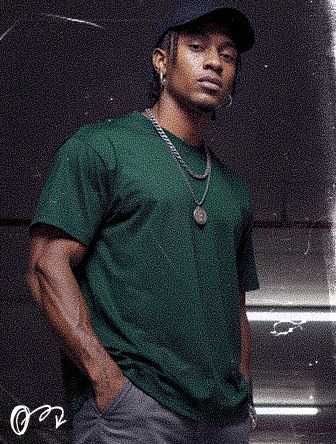Man wearing a green shirt and cap, standing confidently with hands in pockets, adorned with a necklace against a dark, textured background.