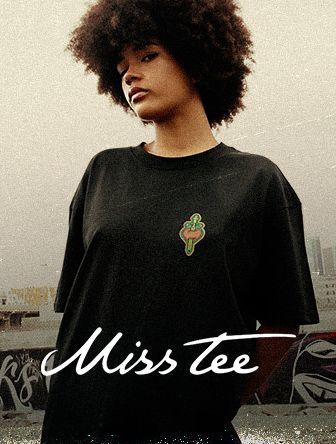 Person with curly hair wearing a black T-shirt featuring a colorful emblem. Text reads "Miss Tee." Urban background.