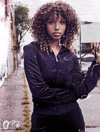 Woman with curly hair in a black zip-up jacket stands confidently on a graffiti-lined urban street, slightly holding the string of her jacket.