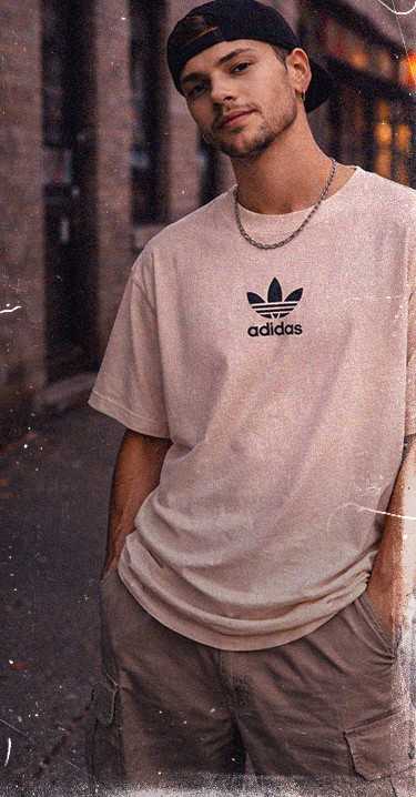 Young man in a light adidas t-shirt and cap stands casually with hands in pockets, in an urban setting with dim lighting.