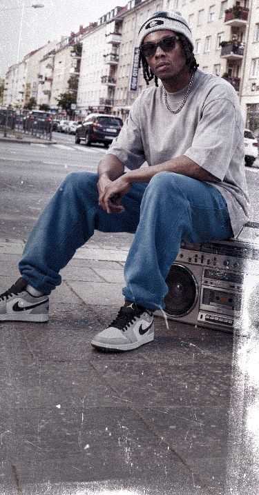 Person in casual streetwear sits on a boombox on a city sidewalk, wearing sunglasses and a beanie. Urban buildings line the background.