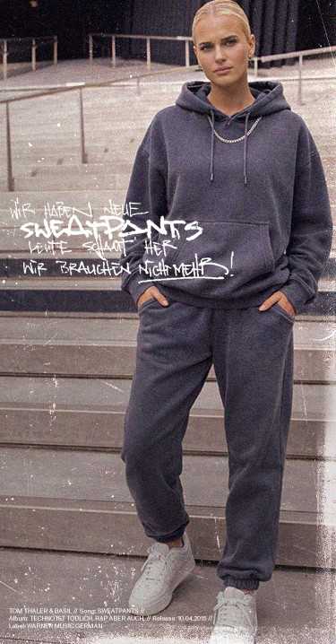 Person in a gray hoodie and sweatpants standing on stairs, hands in pockets, with white sneakers, against a graffiti-style text wall.