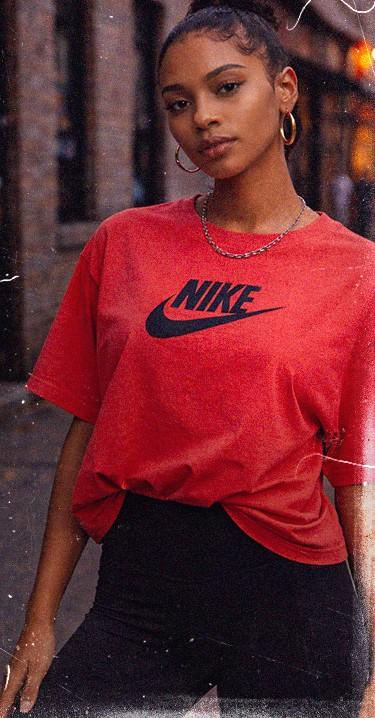 A person in a red Nike shirt and black pants stands confidently outside, wearing hoop earrings and a chain necklace.