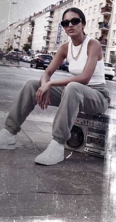 Woman in sunglasses and casual outfit sitting on a boombox, in an urban setting with buildings in the background.