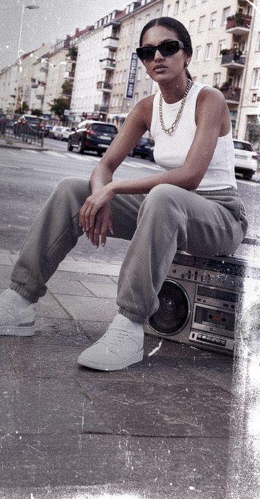 Woman in sunglasses and casual outfit sitting on a boombox, in an urban setting with buildings in the background.