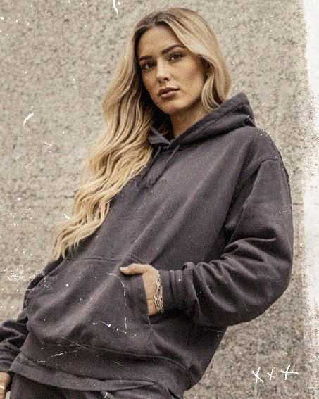 Woman with long blond hair in a dark hoodie stands against a textured wall, looking confidently at the camera.