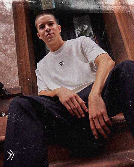 Person sitting on steps, wearing a white T-shirt and dark pants, looking confidently at the camera with a slight tilt.