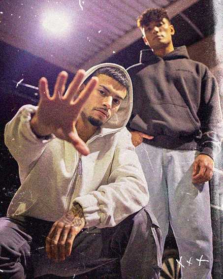 Two men posing confidently; one crouches with his hand extended, wearing a white hoodie, while the other stands behind in a black hoodie.