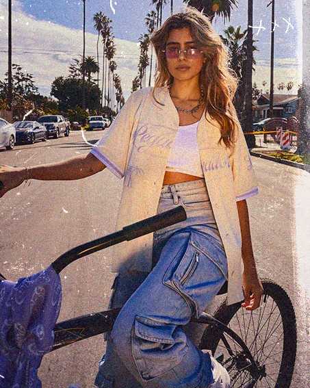 Fashionable person with sunglasses standing beside a bicycle on a sunny street lined with palm trees.