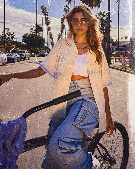 Fashionable person with sunglasses standing beside a bicycle on a sunny street lined with palm trees.