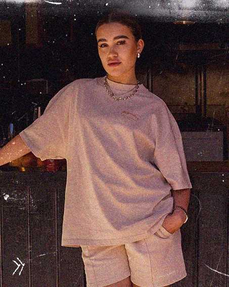 Woman in casual beige outfit leans on a counter, with one hand in her pocket and a confident expression, in dim lighting.
