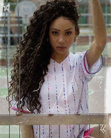 Woman with long curly hair, in a white and striped shirt, stands behind a metal fence, gazing directly at the camera.