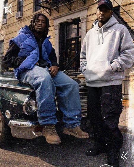 Two individuals pose in casual attire on a vintage car in an urban setting, one sitting and the other standing, both looking at the camera.