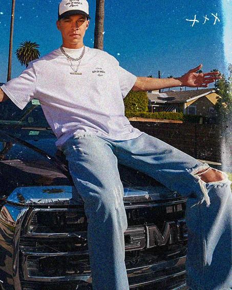 A person in a white cap and T-shirt sits on a GMC truck hood, wearing ripped jeans. Palm trees and houses are visible in the background.