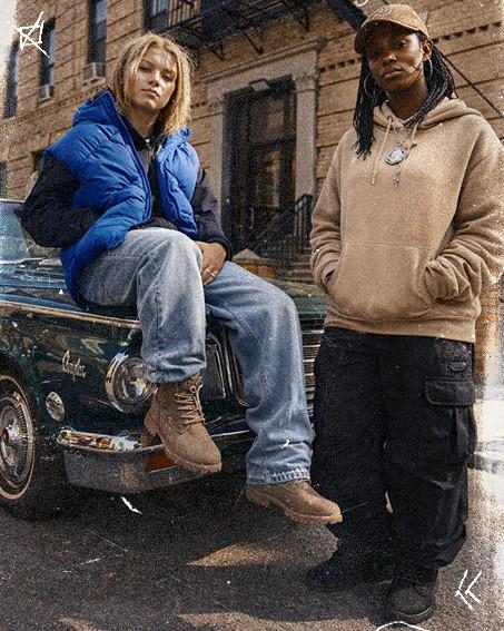 Two individuals in casual outfits pose in front of a vintage car on a street. One sits on the hood, the other stands beside it.