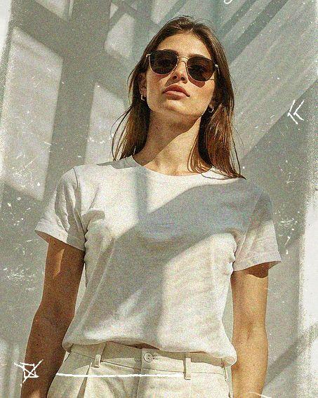 Woman in sunglasses and white t-shirt stands confidently under dappled sunlight, with geometric shadows in the background.