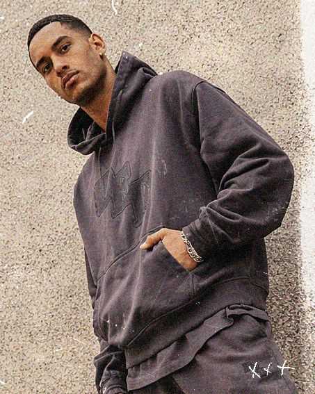 Man in a dark hoodie and pants stands against a textured concrete wall, looking at the camera with one hand in his pocket.