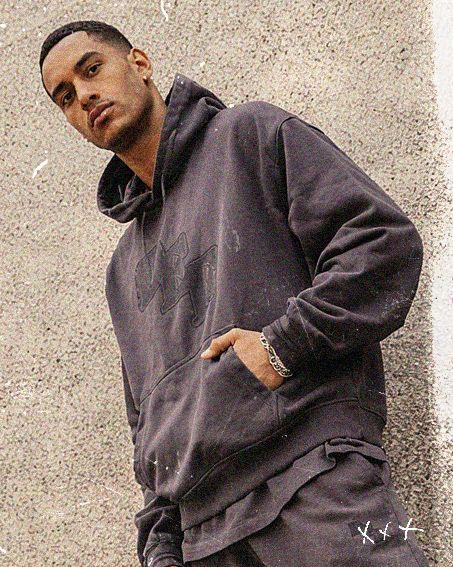 Man in a dark hoodie and pants stands against a textured concrete wall, looking at the camera with one hand in his pocket.