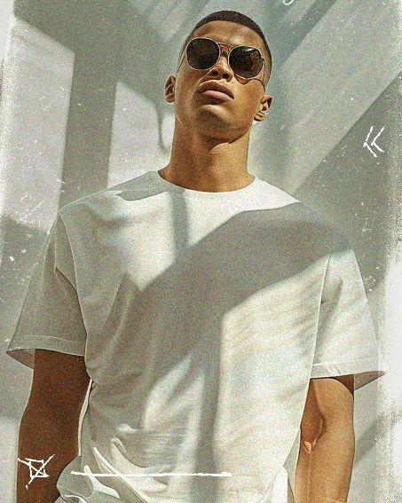 Man in sunglasses wearing a white t-shirt, standing in dappled sunlight with shadow patterns on the wall behind him.