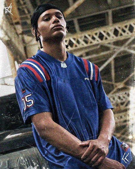Person in a blue sports jersey leans against a vehicle under a metal bridge, with arms crossed and a focused expression.