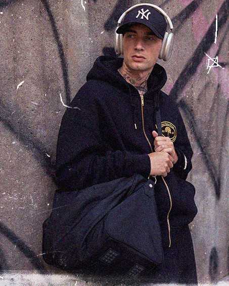 Person in a hoodie and cap, wearing headphones, standing against a graffiti-covered wall, holding a black bag.