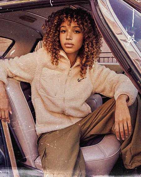Person with curly hair sitting in a car wearing a beige fleece jacket and olive pants, with one arm resting on the open door.