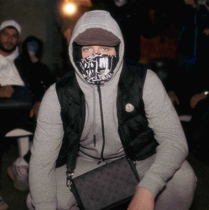 Mann hockt auf dem Boden mit in einem Zipphoodie, Weste und Louis Vuitton Tasche