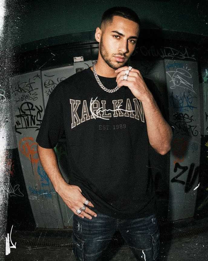 Man in a black "Karl Kani" shirt poses in front of graffiti-covered walls, wearing a chain necklace and rings, with a thoughtful expression.