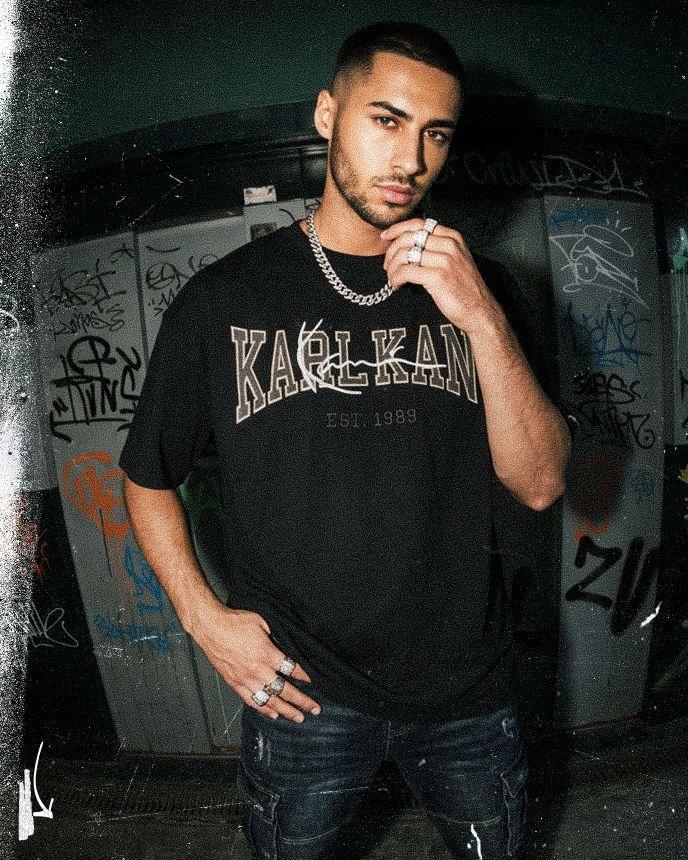 Man in a black "Karl Kani" shirt poses in front of graffiti-covered walls, wearing a chain necklace and rings, with a thoughtful expression.