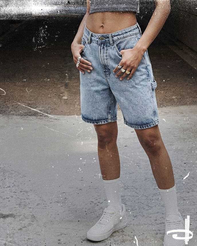 Person in a cropped gray top and knee-length denim shorts, wearing white sneakers and socks, stands on a textured surface with hands in pockets.