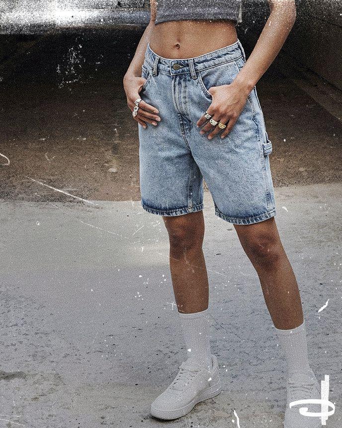 Person in a cropped gray top and knee-length denim shorts, wearing white sneakers and socks, stands on a textured surface with hands in pockets.