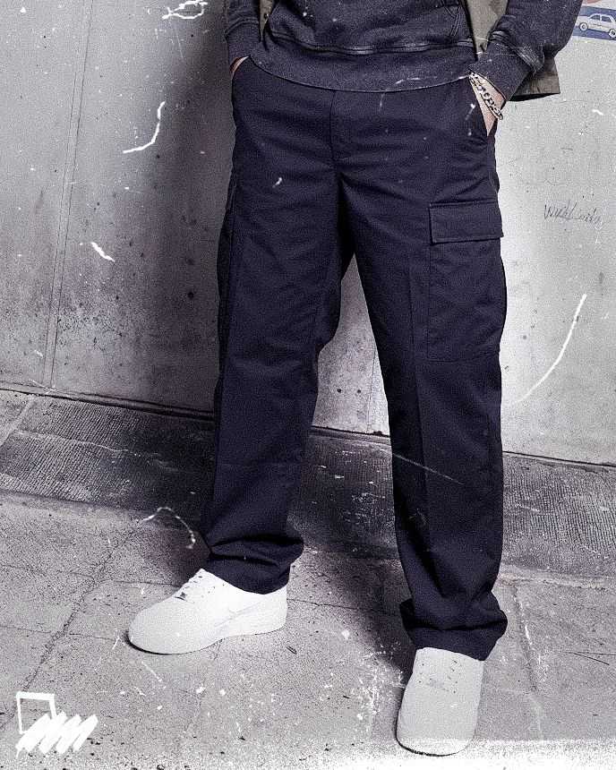 Person wearing dark cargo pants, white sneakers, and a sweatshirt with hands in pockets, standing against a textured concrete wall.
