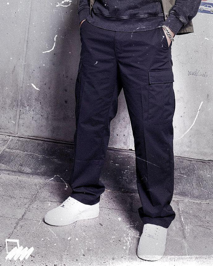 Person wearing dark cargo pants, white sneakers, and a sweatshirt with hands in pockets, standing against a textured concrete wall.