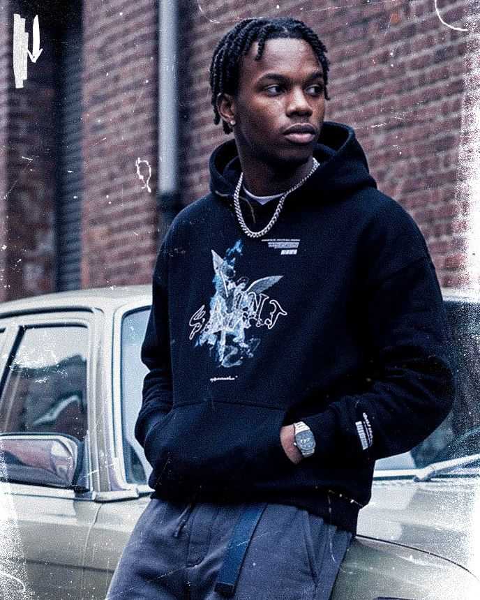 Man in black hoodie and chain leans on a vintage car, looking away, with a brick wall background.