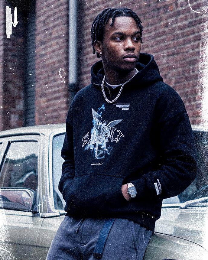 Man in black hoodie and chain leans on a vintage car, looking away, with a brick wall background.