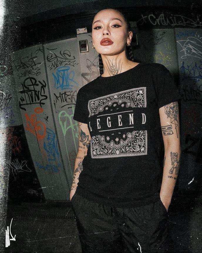 Person with tattoos wearing a "Legend" T-shirt stands confidently in front of a graffiti-covered wall.