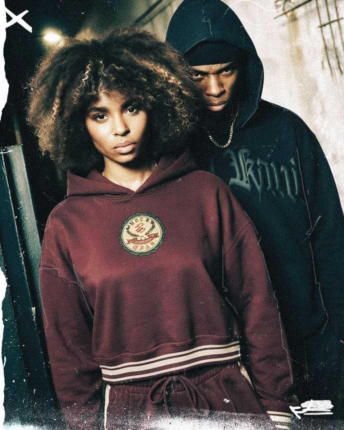 Two individuals standing in an urban setting, one with curly hair in a maroon hoodie and the other behind in a dark hoodie, looking serious.