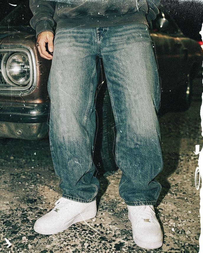 Person wearing faded jeans and white sneakers stands next to a vintage car on a gravel surface.