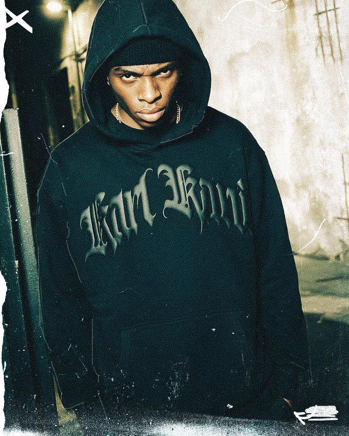 A person wearing a dark hoodie with "Karl Kani" lettering stands in a dimly lit urban alley, looking directly at the camera with a serious expression.